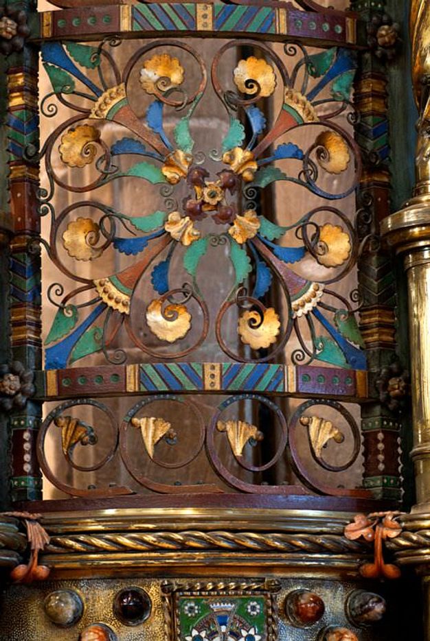 Architectural detail. Panel with ironwork wrought into a four-leaf, double-lobed design, like a lucky clover, is set into a square frame. It and the surrounding ornament are gold-coloured or painted brilliant blue, muted purple, pine green, or umber orange.