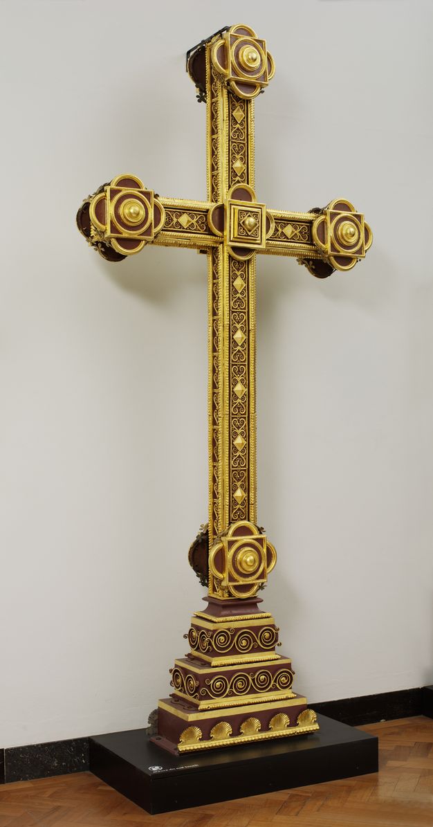 Sculpture. A cross standing on a three-stepped base is decorated with shiny gold geometric designs against a brick-red background.