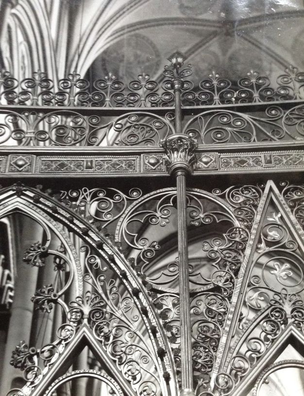 Choir screen. This black and white photograph focuses on the intricately swirling ironwork along the top edge of a choir screen. The vaulted ceiling of the church is glimpsed above.