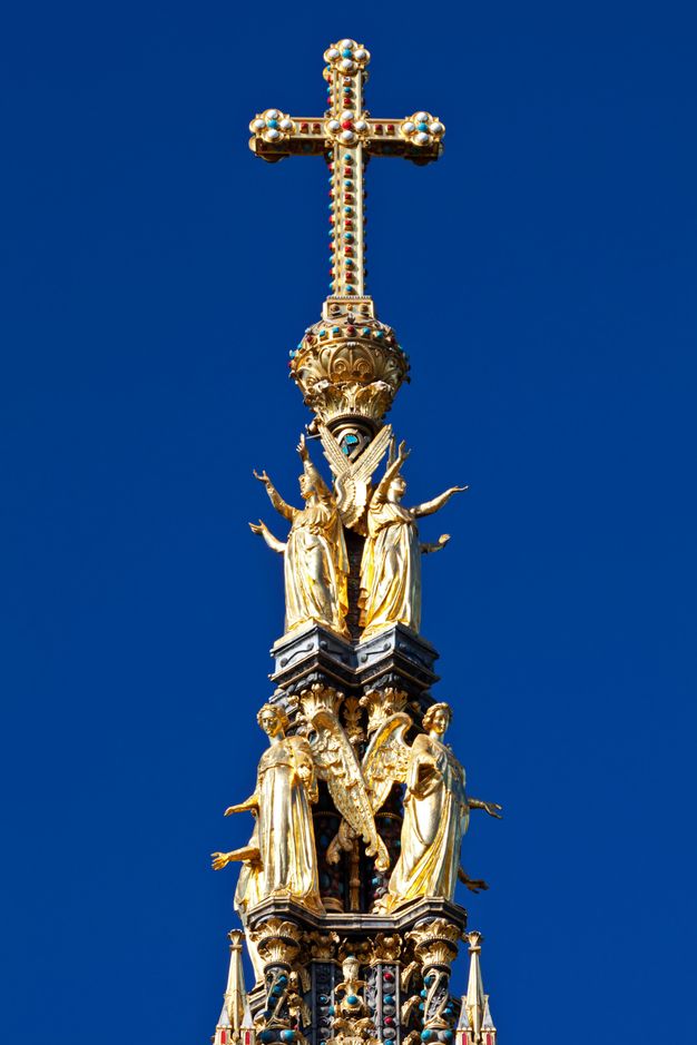 Architecture. A gold, jewelled cross sits on a base held up by two rows of gilded, winged angels against an ultramarine-blue sky.