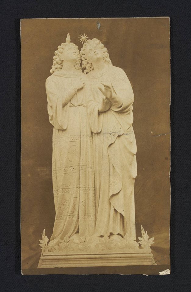 Sculpture. Printed in sepia-brown tones, this photograph shows a pair of statues sharing a base. They both have shoulder-length curly hair, wear long robes, and look up. One has a flickering flame over the head and the other a starburst.