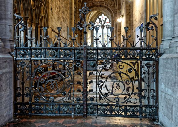 Metalwork. Black iron gates decorated with swirling and vegetal patterns block access to the side aisle of a church. A greenish patina is visible on their surfaces.