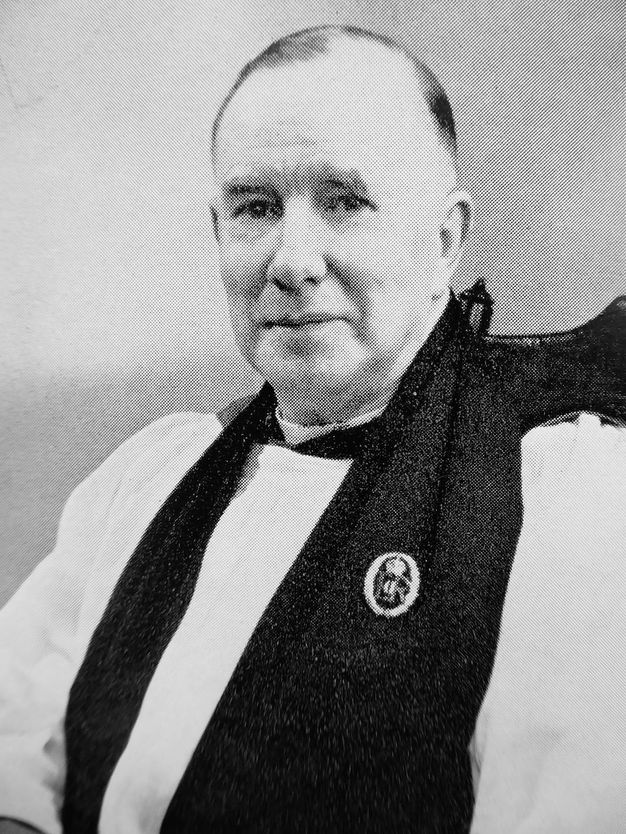 Black and white photograph. Shown from the chest up, a pale-skinned man looks at us with dark eyes under bushy brows. He is clean shaven with sagging jowls. He wears clerical robes and sits in front of a blank background.