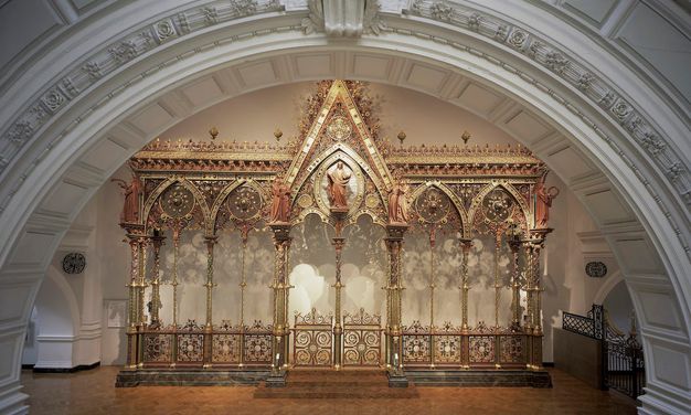 Choir screen. Freestanding architectural partition, like a floating wall, has five bays, each under a pointed arch. Each arch is then bisected by a spindly column. The central bay is the widest and tallest, and a person stands at its apex. Intricate tracery encrusts the upper edges of the screen and fills in the arches. Panels across the bottom are also made with intricately patterned tracery. Most surfaces are shiny or burnished gold, and there are also touches of muted red, warm yellow, and earthy green.