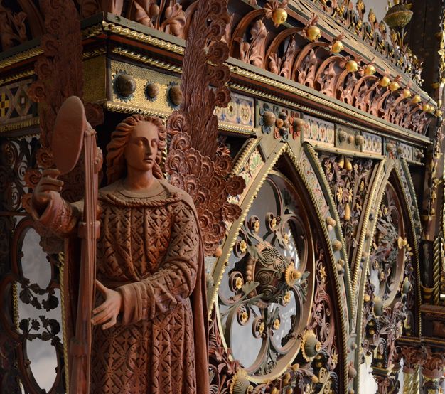 Choir screen. Colour photograph with detail view of a muted red winged angel holding a large standing instrument against intricately painted tracery.
