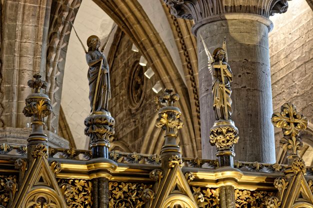 Sculpture. Perched high on an altarpiece, two winged angels carved from stone each hold a cross and a rod. The objects, their clothing, halos, and ornamental details below are gilded.