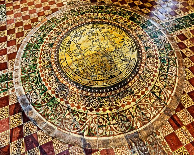 Pavement. A roundel is set within a diamond checkerboard pattern made up of russet-red and floral-patterned tiles. In black outlines, the scene in the roundel shows a man behind restrained by two others, as an executioner raises a sword. Seven concentric rings of pattern telescope out from the roundel.