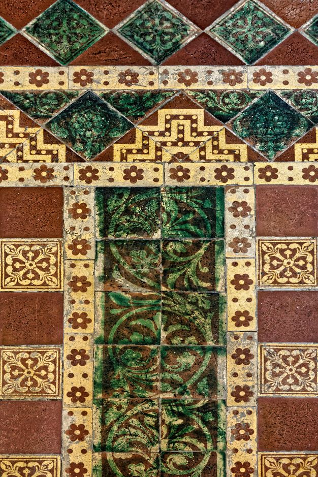 Pavement. A detail shows forest-green tiles decorated with curling vines, set into a framework of longer, thin tiles with rosettes against a cream-white background. Other tiles, in dark red or floral-patterned, create an intricate design.