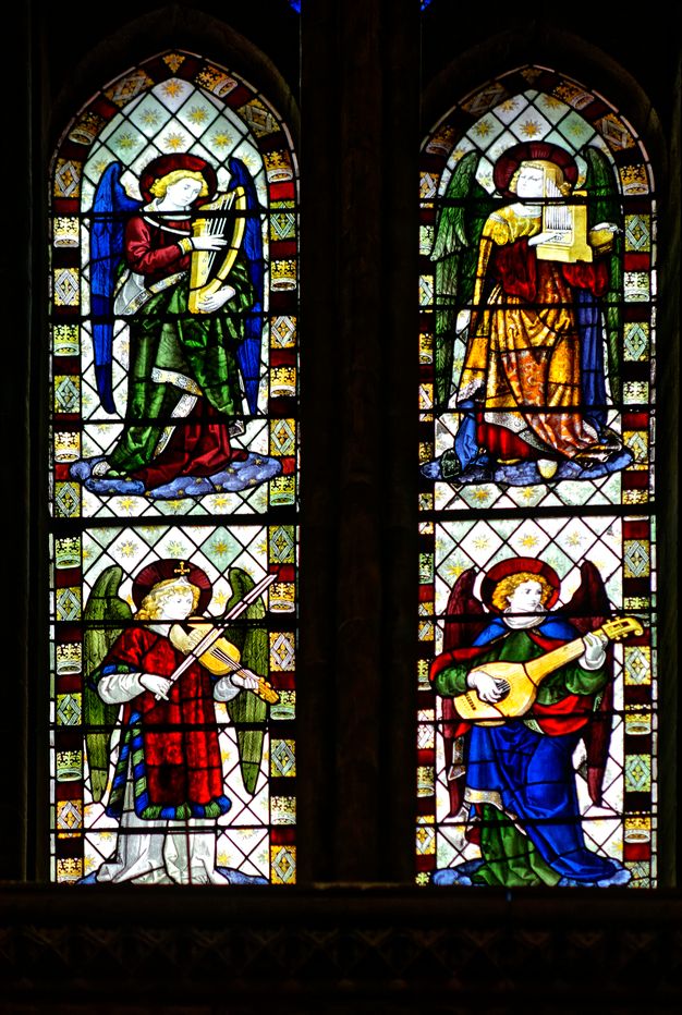 Stained glass window. Two pointed arched windows each show two winged angels playing musical instruments, one over the other, for a total of four. The clothing and objects are created with lapis-blue, emerald-green, crimson-red, and butterscotch yellow glass.