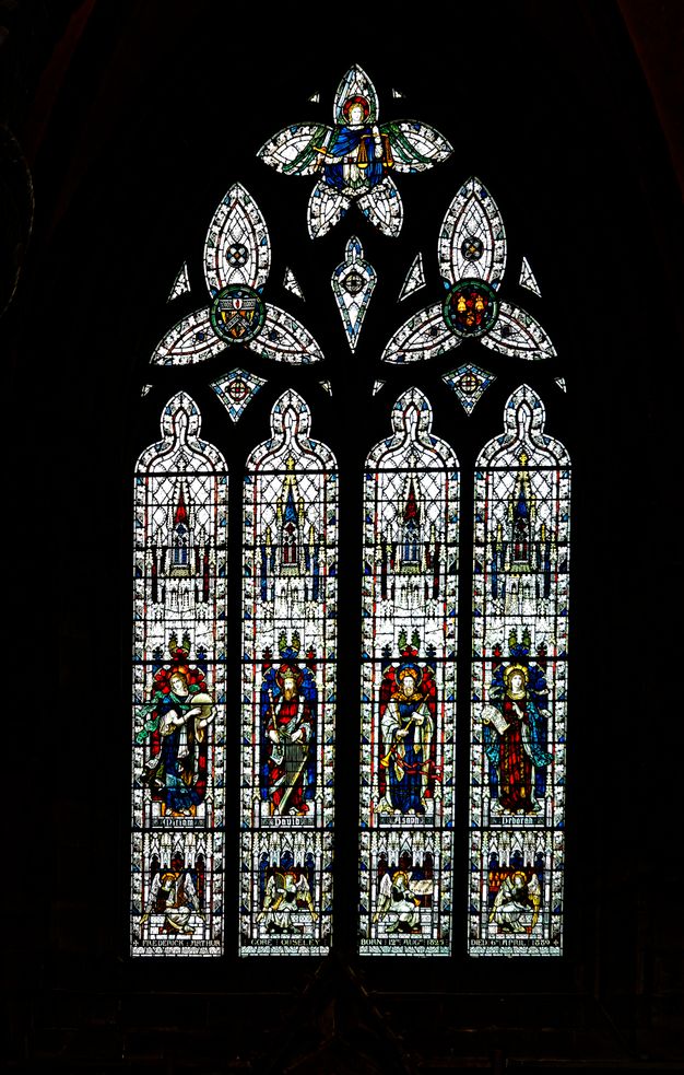 Stained glass window. Four people stand, each in a tall narrow pane, and each holds a musical instrument. The four panels are topped with trefoil and five-lobed windows. Flecks and slivers of glass between the larger components glint like sparks against the black surround.