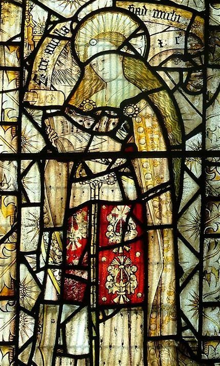 Stained glass window. Detail view of a haloed woman with long blond hair, the Virgin Mary, reading a book. Pieces of glass around her have sunbursts overlaid with blooming white roses.