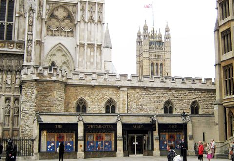 Architecture. A two-storeyed, hall-like stone structure stretches across a church façade. The second level has two-part, pointed arched windows and crenelation along the roofline. The lowest level has storefront windows and the sign, “The Westminster Abbey Shop”.