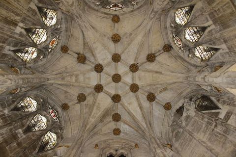 Architecture. We look up at a ribbed grey stone ceiling which has a hexagonal shape and stained glass windows on several sides. Knots of decorative foliage sit at each site where the ribs intersect.