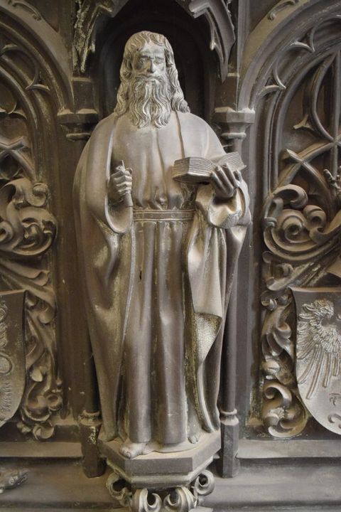 Sculpture. A man with a long beard and hair, and wearing robes belted at the waist, stands holding an open book in one hand. He holds another object in his other hand, perhaps a scroll. The hexagonal platform on which he stands is supported on leafy flourishes. The surfaces are muted, silvery gold.