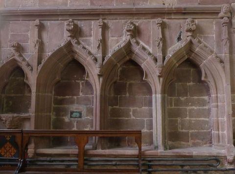 Architecture. Four niches under pointed arches are backed by pale red stone. The arch over each niche comes to a point and is topped with a finial. The niche to our left is smaller, perhaps two thirds, the size of the others.