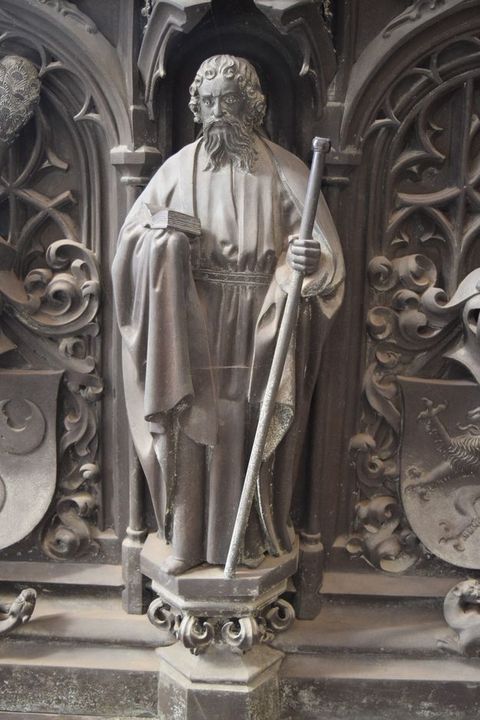 Sculpture. A man with curly hair and a long, forked beard wears robes belted at the waist. He holds an open book in one hand and a staff with a knob at the top in the other. The hexagonal platform on which he stands is supported on leafy flourishes. The surfaces are muted, silvery gold.