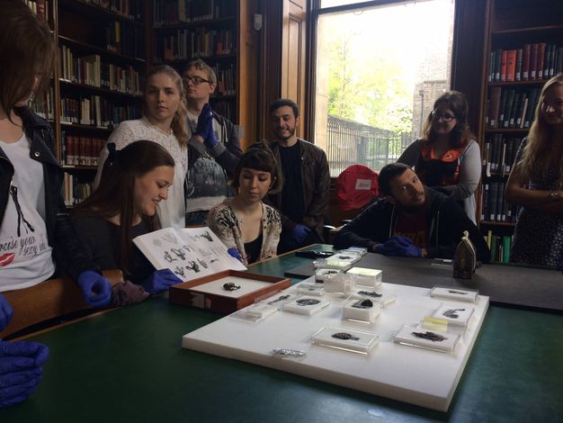 Colour photograph: Nine men and women gather around a table laid with artefacts in a book-lined room. Many look at a book held open by a woman sitting at the table. She and others wear blue gloves.