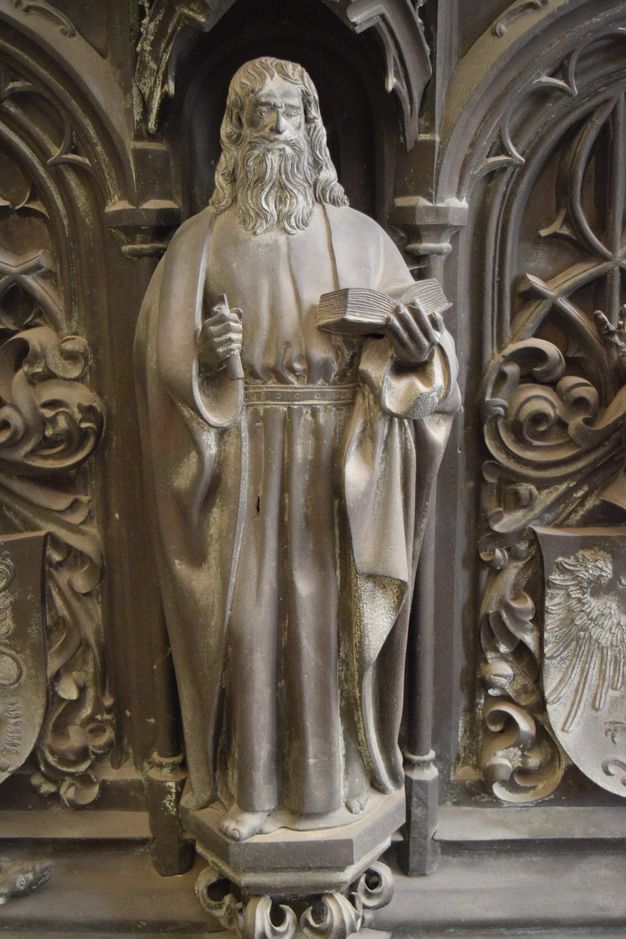 Sculpture. A man with a long beard and hair, and wearing robes belted at the waist, stands holding an open book in one hand. He holds another object in his other hand, perhaps a scroll. The hexagonal platform on which he stands is supported on leafy flourishes. The surfaces are muted, silvery gold.
