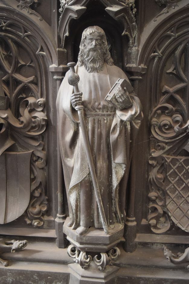 Sculpture. A man with a long beard and hair, and wearing robes belted at the waist, stands holding a closed book in one hand. In the other, he holds a staff with a knob at the top and a large shell below it. The hexagonal platform on which he stands is supported on leafy flourishes. The surfaces are muted, silvery gold.