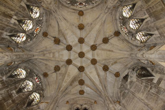 Architecture. We look up at a ribbed grey stone ceiling which has a hexagonal shape and stained glass windows on several sides. Knots of decorative foliage sit at each site where the ribs intersect.