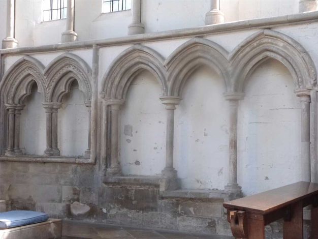 Architecture. Three pointed arches outlined in grey stone against a white wall create three niche-like spaces. A wide ledge below steps down in each successive niche, resulting in seats at three levels.