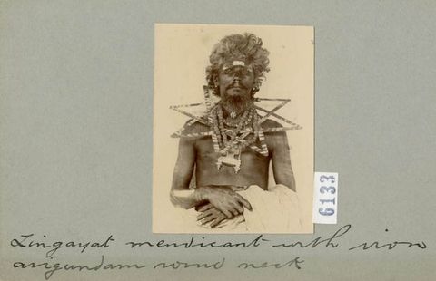 Black and white photograph. Shown from the waist up, a dark-skinned man sits looking at us with his hands clasped in his lap. Flat triangular or star-shaped forms lie around his neck and across his shoulders like stiff, oversized necklaces. The photograph is mounted on a card with handwritten text beneath.