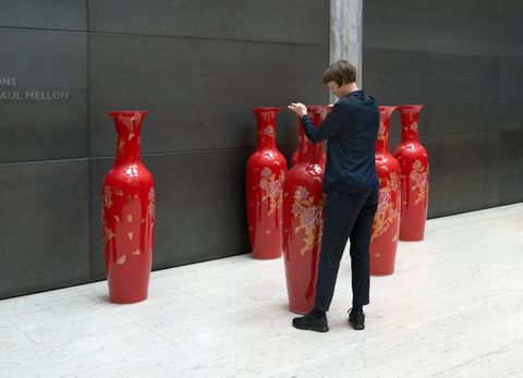 Installation photograph. A woman adjusts the placement of a slender vibrant red vase that comes up to her shoulder height. Five more vases are along or near a black wall opposite us.