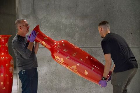 Two men carry a slender, vibrant red vessel. Both wear purple gloves. One supports the vessel by the foot, the other around the neck. The vessel is nearly as tall as they are, and they shift its position in front of a grey wall.