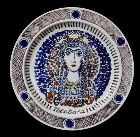 Plate. A woman’s head and shoulders are sketchily painted mostly in shades of blue and brown and with a few touches yellow. Circles over her head and down the sides of her face suggest a headdress and other jewelry. Her name is painted below, \“Theodora\”.
