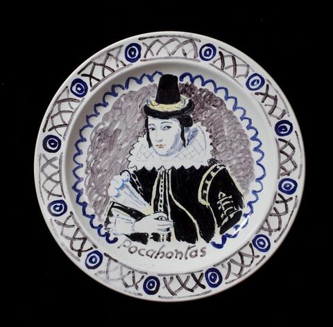 Plate. A woman wearing a black dress, a stiff white collar, and a tall black hat is shown from the waist up. She holds a feather fan in one hand. She is painted mostly in shades of blue, grey, and black, with a few touches of pale yellow. Her name is painted below, \“Pocahontas\”.