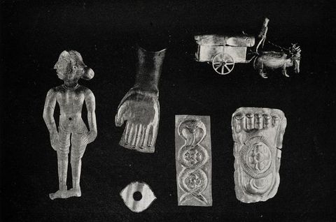 Black and white photograph. Six metal charms are shown against a black background: a woman, a hand, an eye, a cobra, a foot, and a carriage pulled by animals.