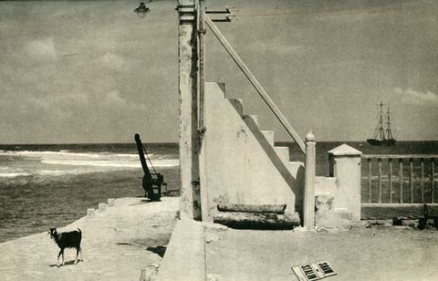 Photogravure. A flight of stairs appears to lead to the top of a telegraph station pole at the centre of this surreal, black and white landscape. A body of water opens beyond the stairs, and on the horizon is a two-masted sailing ship. A goat strolls along a walkway to our left. The cover of a magazine on the ground near us in the lower right corner reads, \“Shaw on Stalin\”.