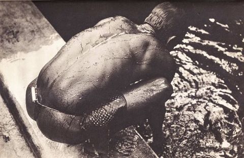 Black and white photograph. The crouching back of a dark-skinned man nearly fills this composition. Water glistens off his skin and runs in a rivulet down the indentation of his spine. He bends forward and reaches into the water beyond the ledge on which he squats.