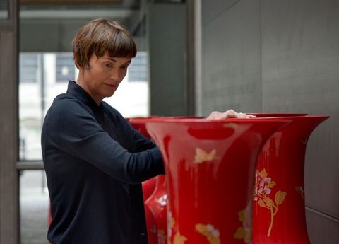 Installation photograph. Shown from the hips up, a woman rests her bare hands on the mouth of a shoulder-high, vibrant red vessel. Her head turns towards us to look at other vessels between her and us.