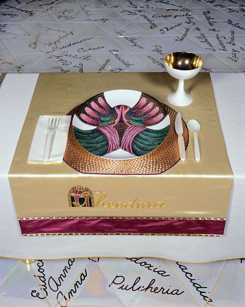Installation. A place setting at a table has a plate painted to look like a mosaic in a gold, magenta, blue, and green. The plate and a gold-lined white goblet sit on a pale gold cloth embroidered in darker gold, reading \“Theodora\”. The capital T is overlaid on an architectural arch.