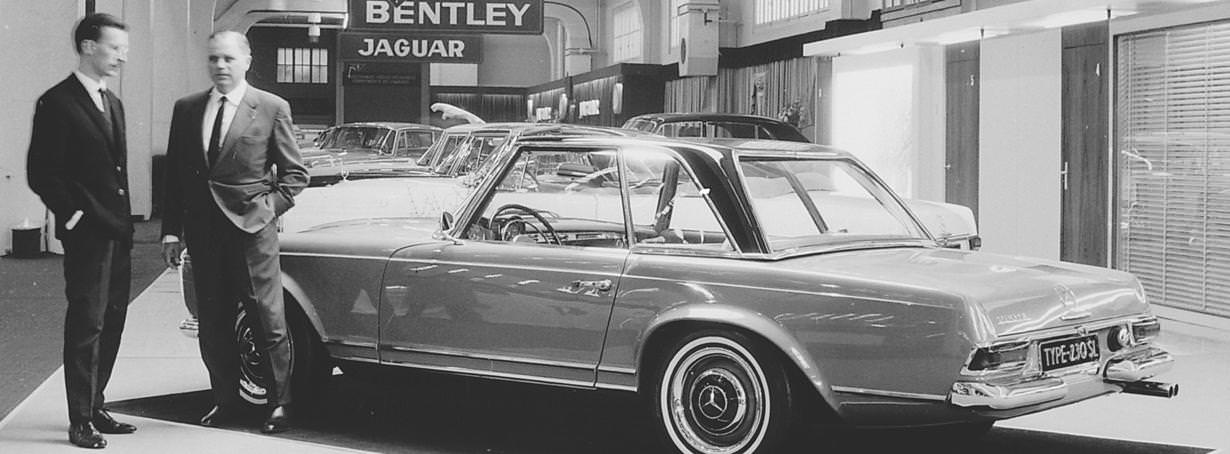 Black and white photograph. Two men wearing suits stand next to a gleaming car in a showroom. Signs further back in the space read \“Bentley\” and \“Jaguar\”.