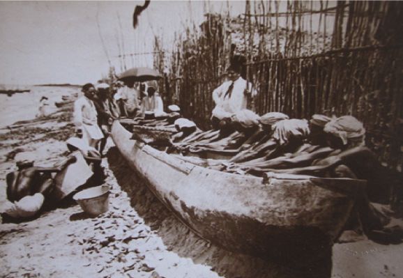 Black and white photograph. On a sandy beach, dark-skinned men with light cloths wrapped around their heads sit alongside a long, wooden boat. Their arms stretch in front of them, engaged in a task hidden by the boat. Men in white clothing look on, some under a parasol at the far end of the boat.
