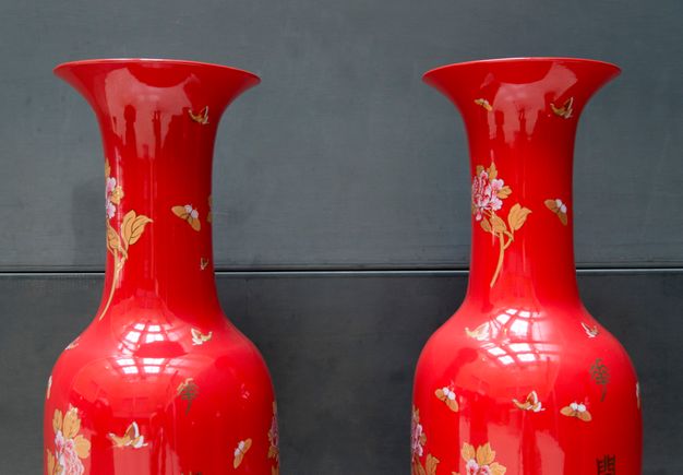 Installation photograph. The vibrant red shoulders, long necks, and flaring mouths of two ceramic vessels reflect the light in the room and the photographer taking this picture. Butterflies and flowers in shades of golden tan, white, and pale pink are scattered across the vessels’ surfaces.