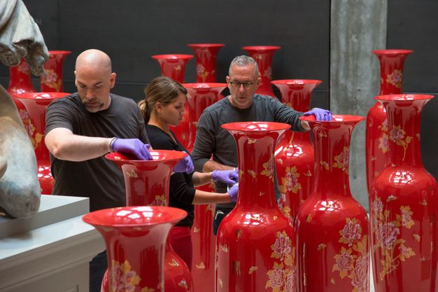 Installation photograph. Two men and a woman are surrounded by fourteen vibrant red ceramic vessels, which come up to chest height on the tallest person. The people wear purple gloves as they touch the openings of the vessels, presumably arranging them in the gallery space.