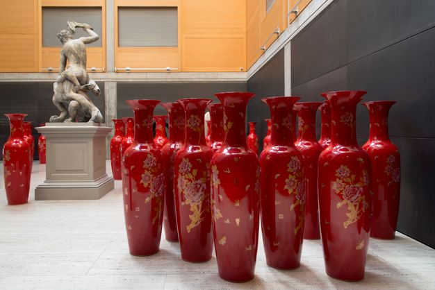 Installation photograph. At least twenty-two vibrant red vessels painted with pink and gold flowers and butterflies stand mostly along one wall in a gallery with stone floors. A sculpture showing two men struggling is on a plinth to our left.