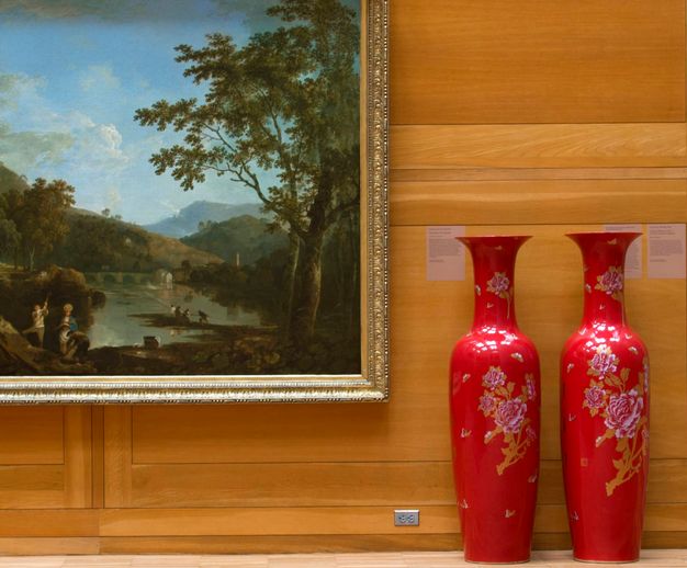 Installation photograph. Two shiny, vibrant red vessels stand in a wood-panelled gallery next to a painting of a vast landscape. The mouths of the vessels come up as high as the wall texts behind them.