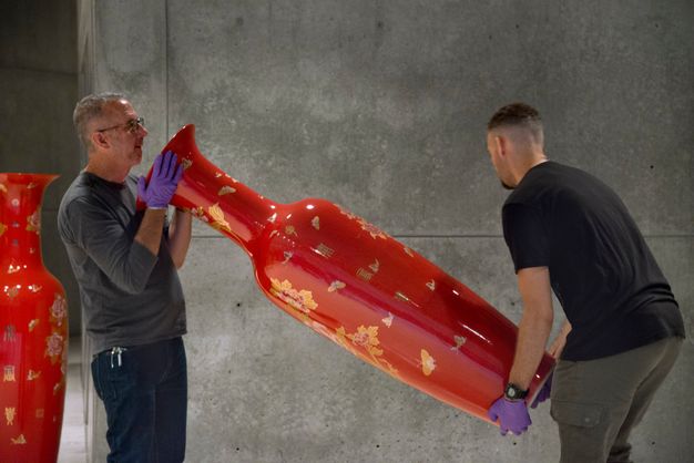 Two men carry a slender, vibrant red vessel. Both wear purple gloves. One supports the vessel by the foot, the other around the neck. The vessel is nearly as tall as they are, and they shift its position in front of a grey wall.