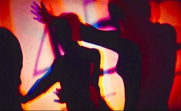 Installation photograph. In this blurred image, dark silhouettes of two people shown from the hips up appear to be dancing in front of a screen on which purple shapes or numbers are being projected. The figures are edged in red, yellow, or blue light.