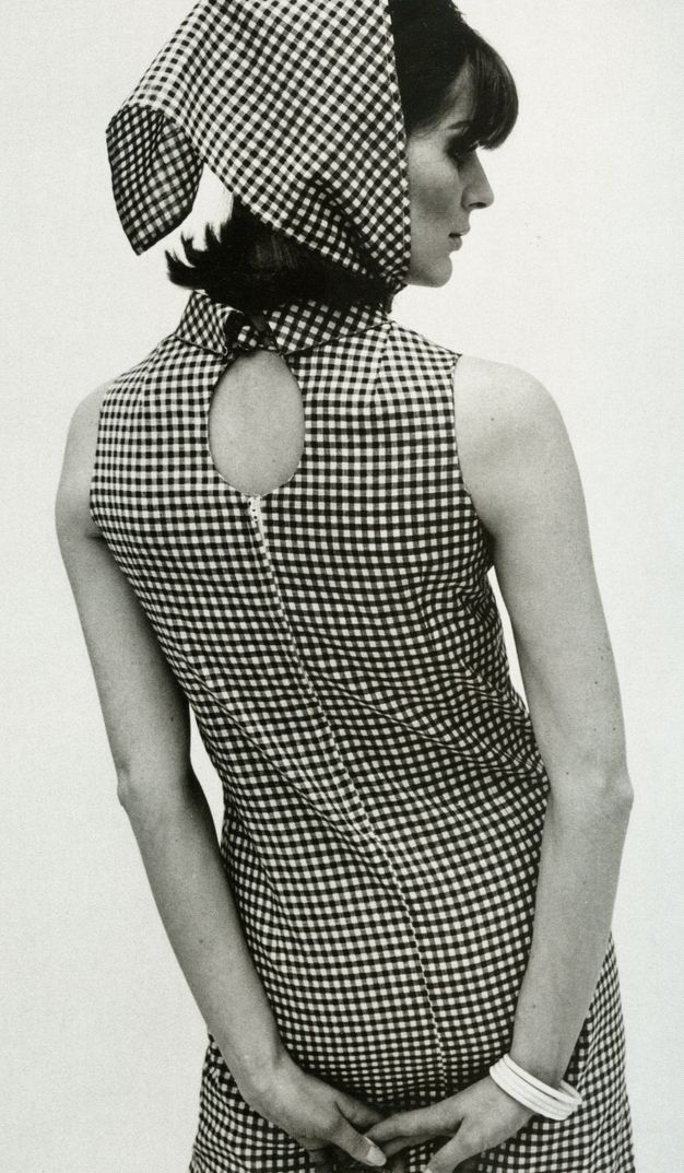 Black and white photograph. Shown from the thighs up, a woman facing away from us wears a checkered sleeveless dress with a keyhole below the high collar. A matching scarf over her dark hair is tied under her chin.