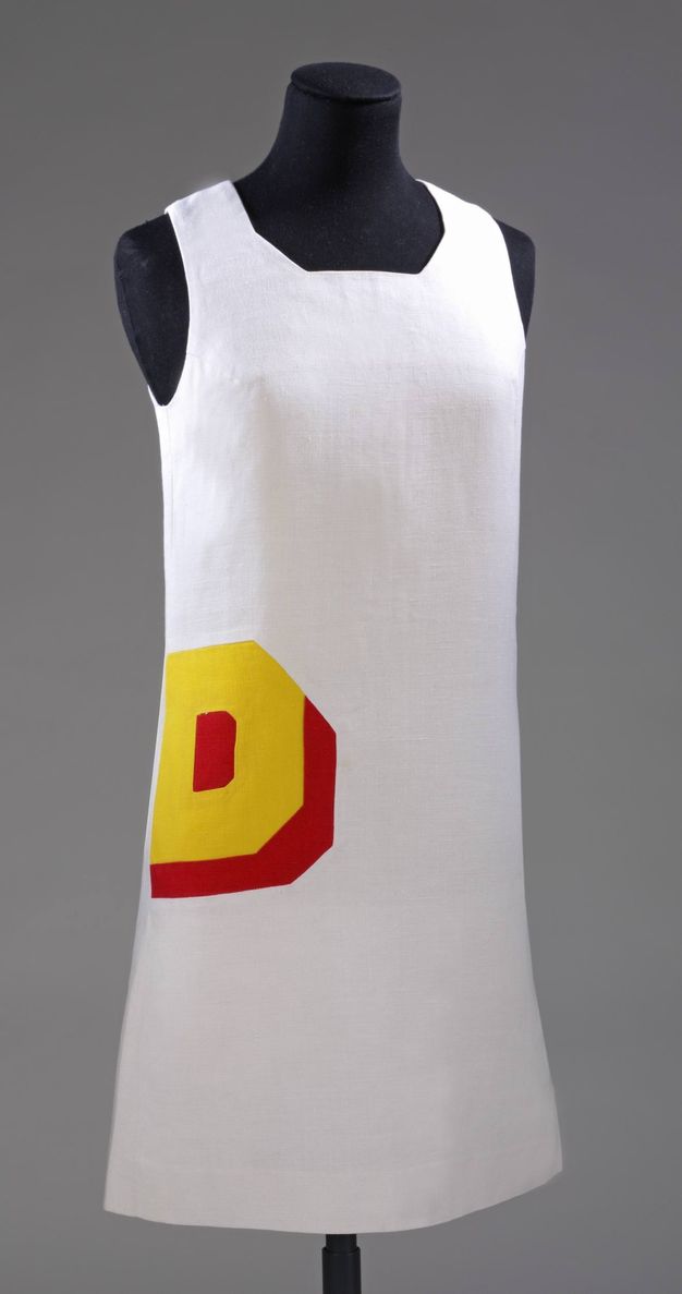 Dress. A white, sleeveless sheath dress on a black tailor's mannequin has a capital letter D in bold yellow, shadowed in scarlet red, over the hip to our left.