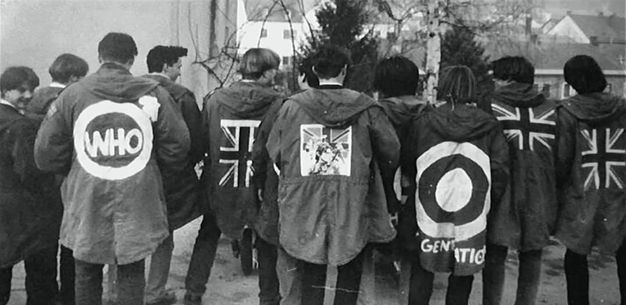Black and white photograph. Eleven young men stand facing away from us to show the backs of their dark, hip-length coats. The Union Jack appears on three jackets. A target with the partially visible word \“Generation\” is on a jacket to our right. A jacket to our left displays the word \“WHO\” in a circle with an arrow pointing diagonally up to our right.