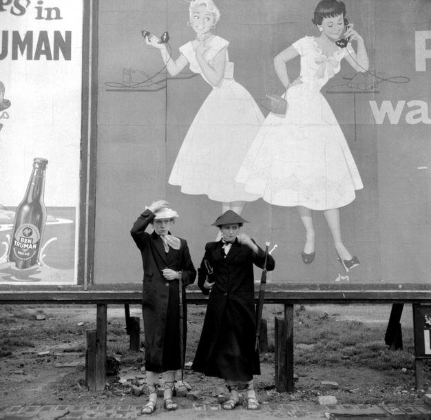 Black and white photograph. Two women wearing long black coats and holding umbrellas hooked over their arms stand in front of a billboard low to the ground. The billboard shows an illustration of a blond and a brunette woman wearing short-sleeved dresses with flaring, shin-length skirts. Each holds an old-fashioned phone receiver.