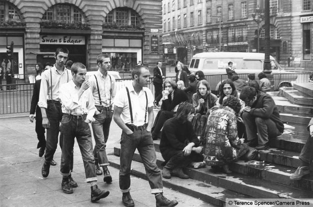 Black and white photograph. Four men with close-cropped hair, white collared shirts, and dark jeans rolled up over boots walk past a group of people sitting on some steps. The men and women in the second group have long hair.