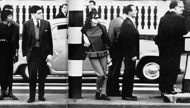 Black and white photograph. Several pale-skinned men in black suits stand along a pavement as a car drives along the street beyond. A woman, also with pale skin, stands leaning on a black and white striped column at the centre of the composition. She wears sunglasses and a shirt with a ruffle along the bottom hem, and she holds what looks like a bottle of milk.