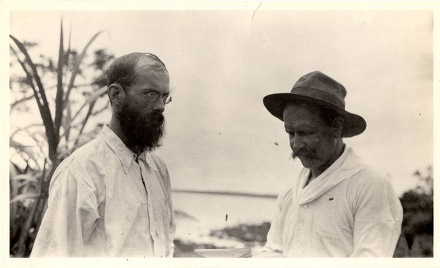 Black and white photograph. Two men are shown from the chest up against a distant sea. Their bodies are angled towards each other as the left man looks off to our right and the other man looks down. Both wear white shirts, and the man on our right wears a fedora.